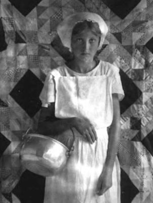 Girl with cap, in front of quilt, holding kettle
