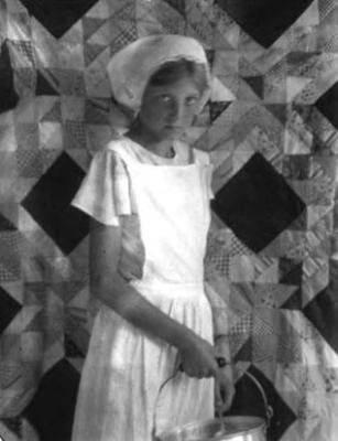 Girl with cap, in front of quilt, holding kettle
